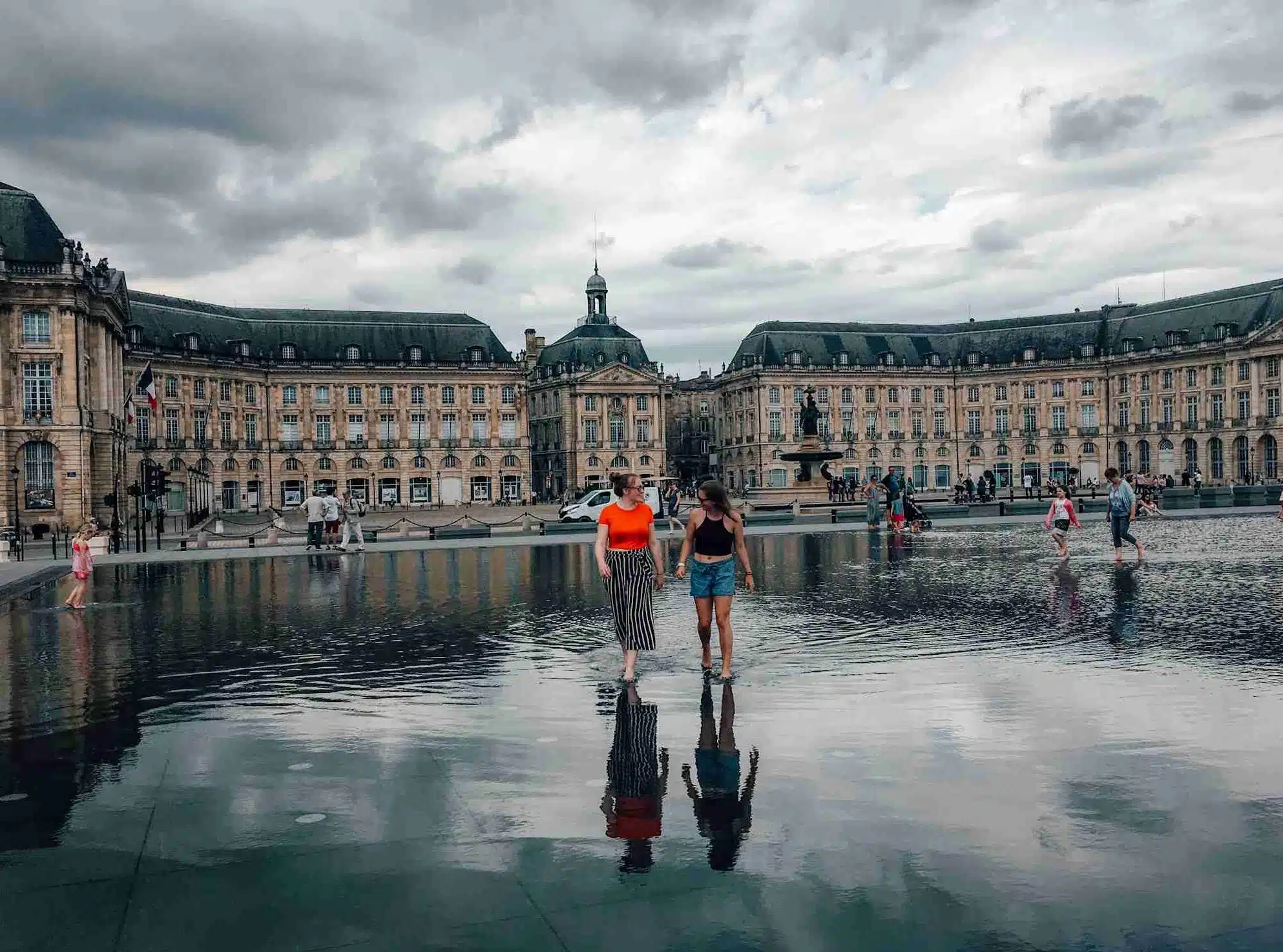 2 girls walking across the Miroir d'eau, a shallow pool of water, on a cloudy day. This is one of the top things to do in Bordeaux