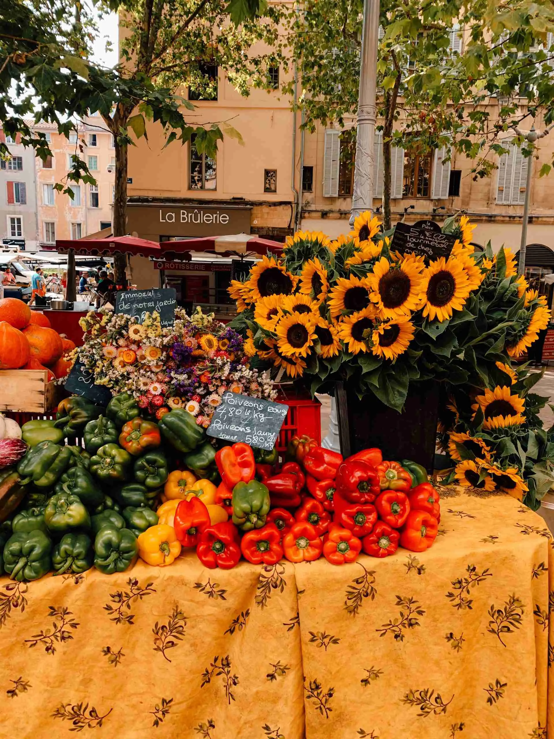 A table full of peppers, flowers and other local product sits on a bright yellow table cloth in the middle of a square in Provence 