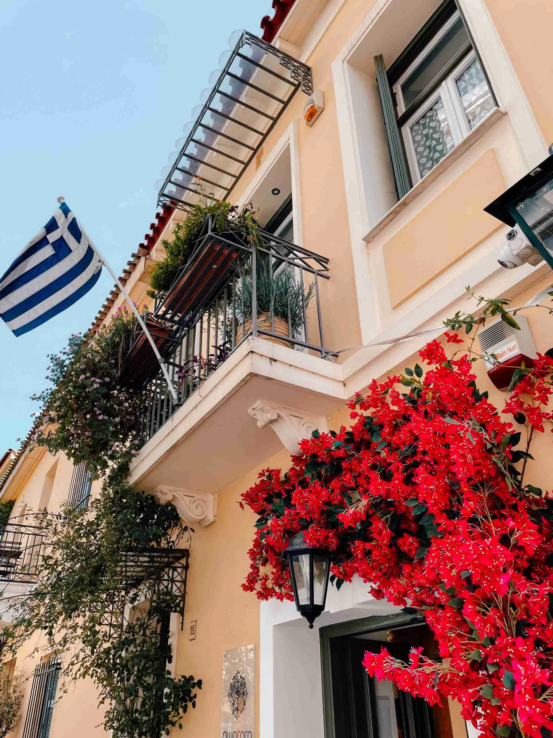 The colourful streets to wander around when visiting Athens. 
A greek flag blows in the wind off of an apartment balcony. Below is a bushel of flowers surrounding the door to the building. 
