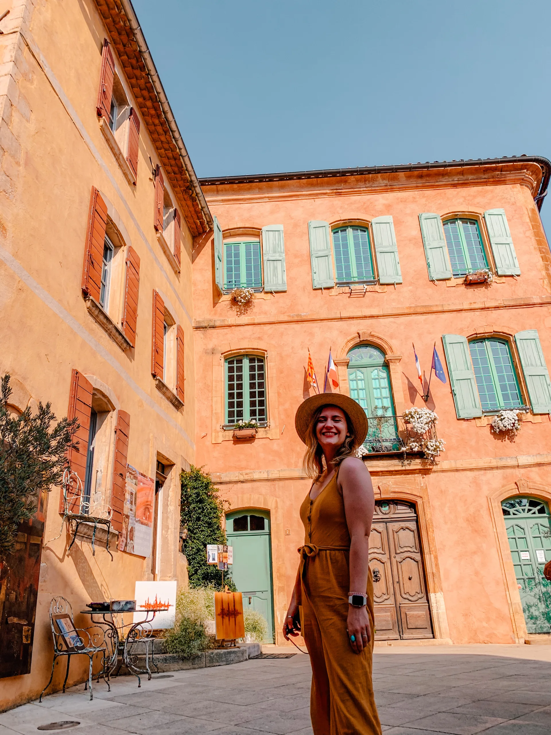 A girl smiles facing the camera with a beautiful orange building behind her - one of the many in Roussillon 