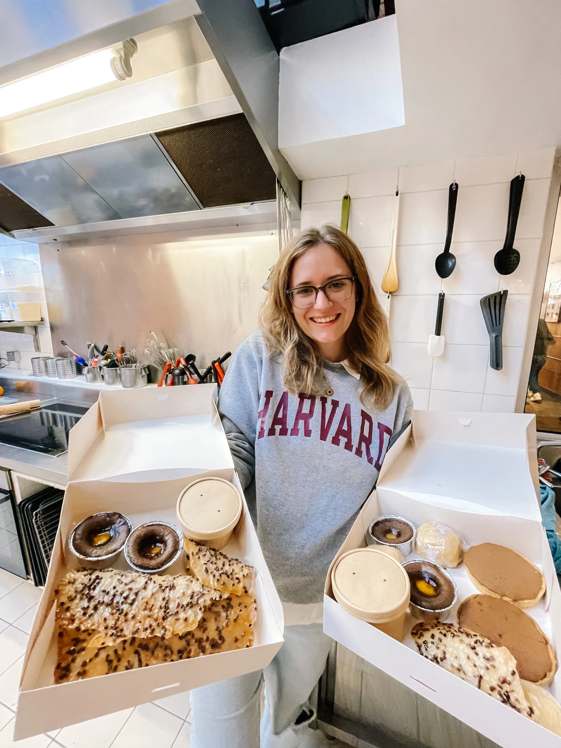 A girl stands holding 2 boxes of freshly baked baked goods in a kitchen 