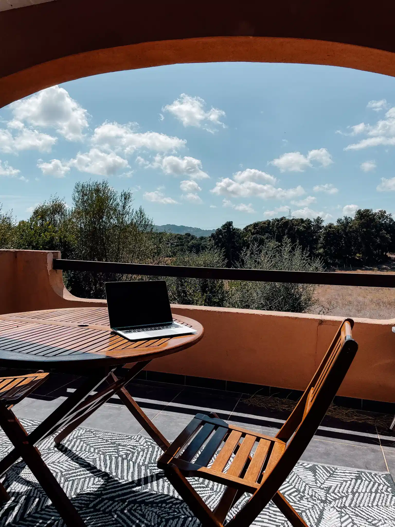 A computer sites on a wooden table on a porch. In the background there is the faint hint of rolling hills with trees and bushes right up to the edge of the porch. 