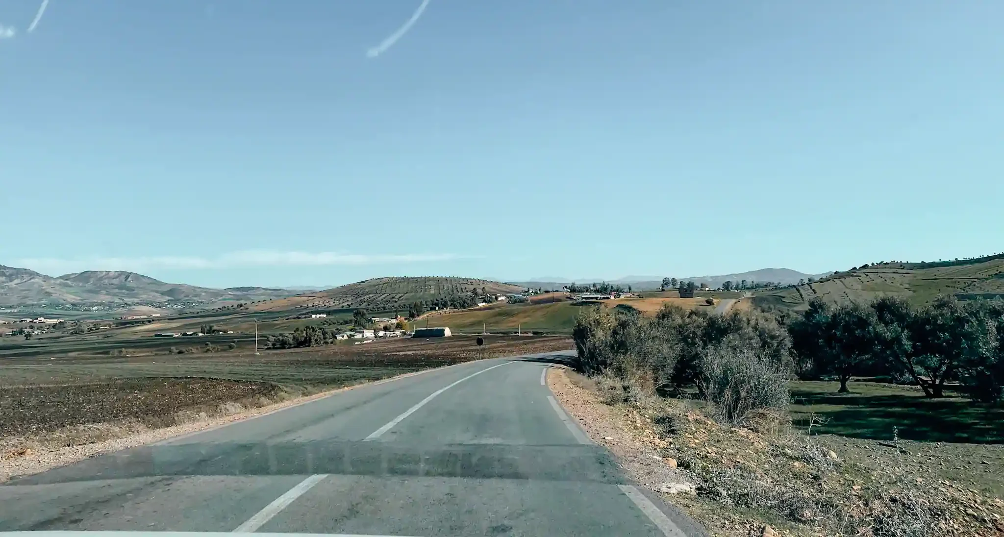 A long highway road in Morocco with rolling hills in the distance and trees lining the road 