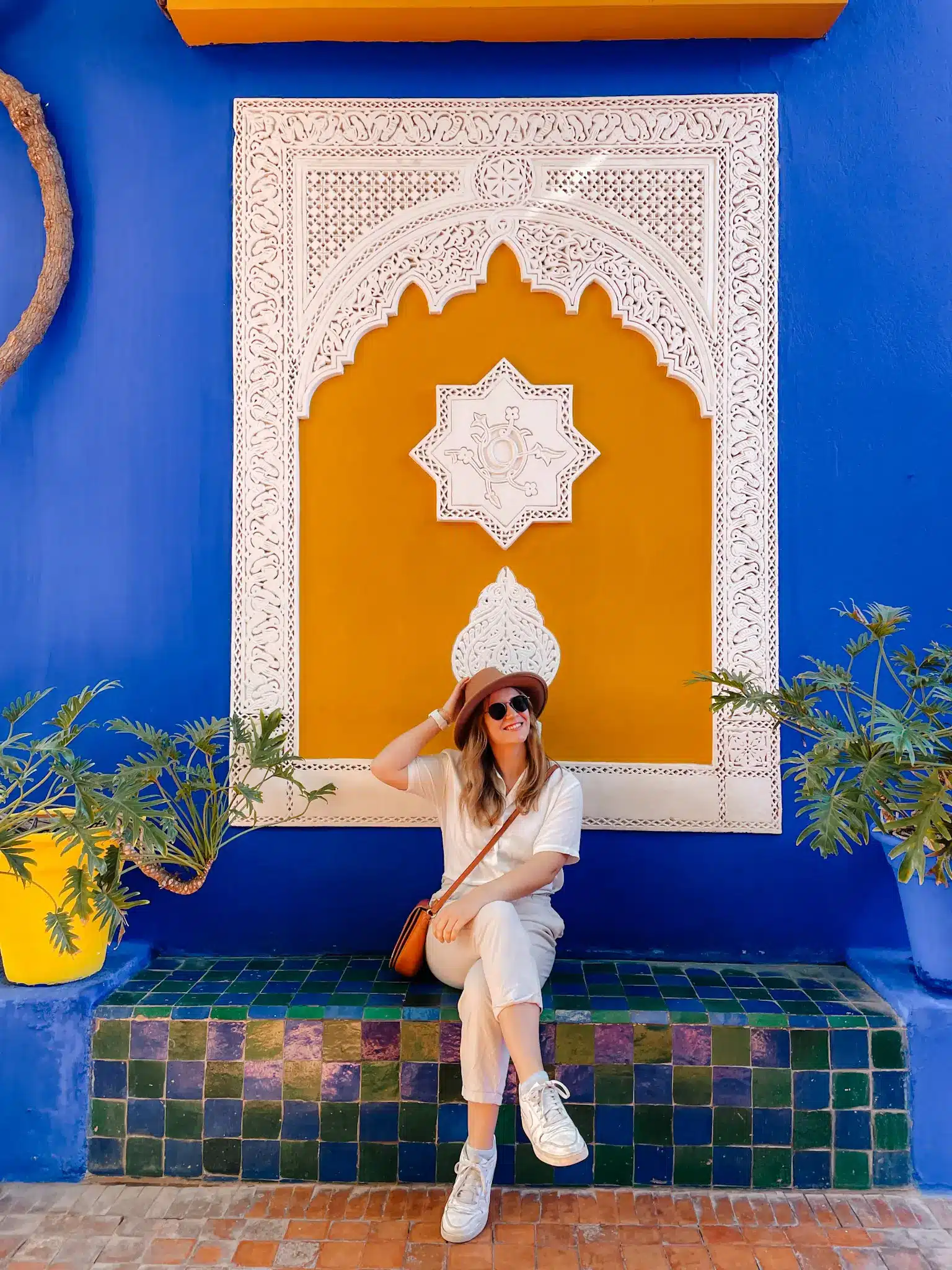 A girl sits crossed legged holding her hat in front of a ornate Moroccan artwork with green plants beside her and a bright blue wall behind her