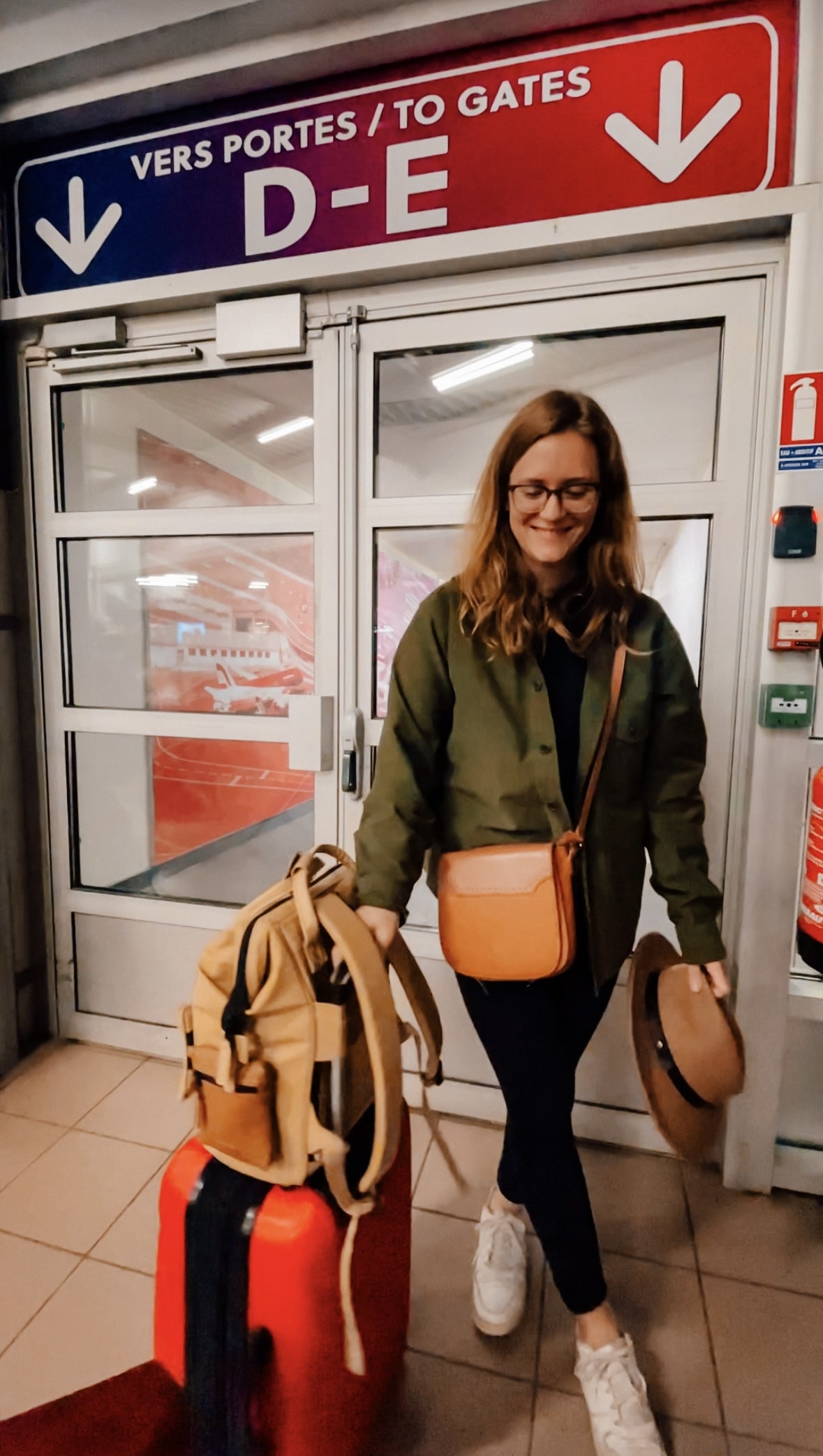 A girl stands next to her boarding gate holding her suitcase with a back pack on top, holding her hat in another hand and a purse wrapped around her shoulders. 