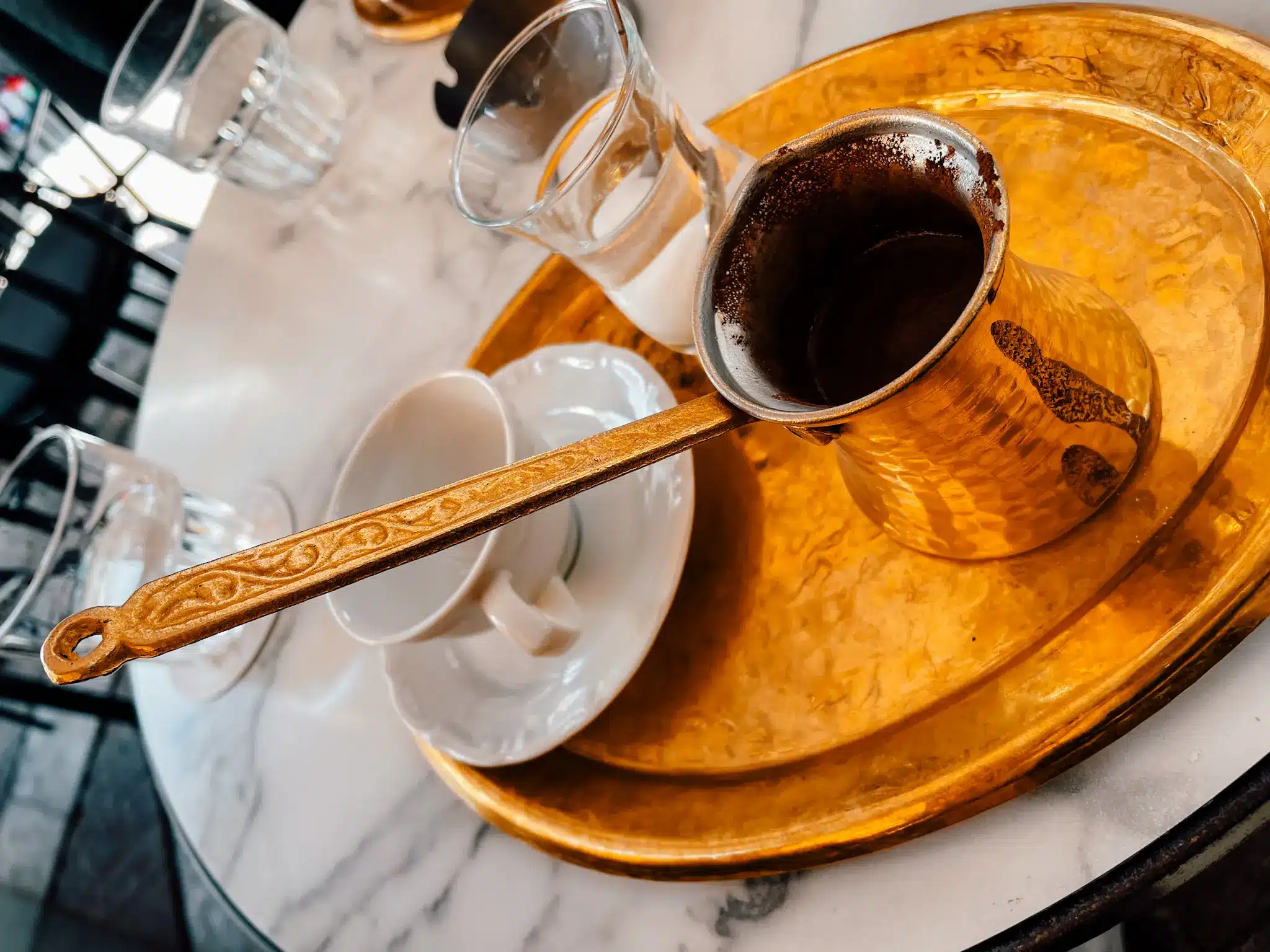 A traditional coffee in Greece sits on a gold plate with a pouring bowl ready to be poured into the coffee cup