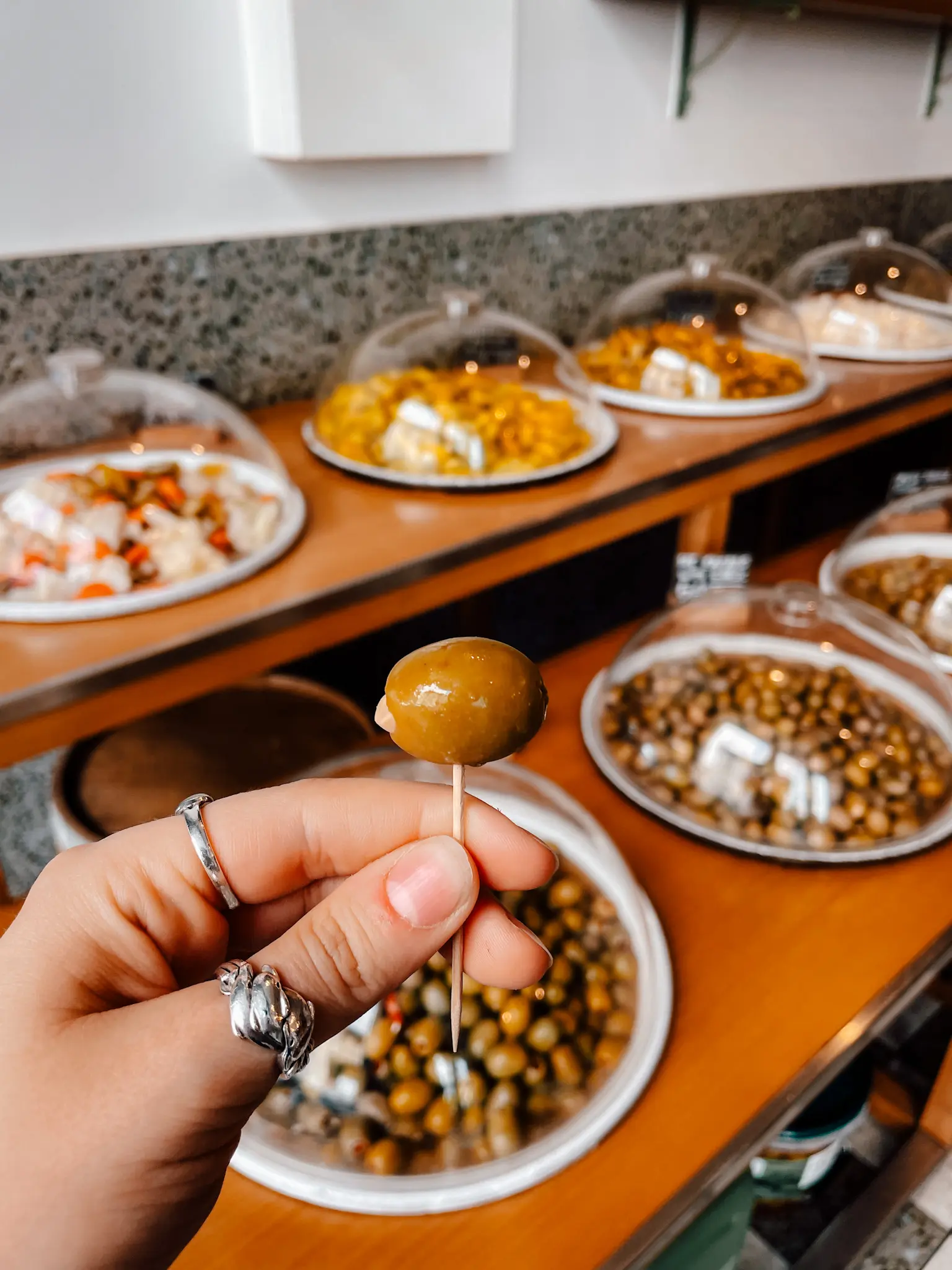 Someone holds an olive on a toothpick with rows of olives in display cases in the background