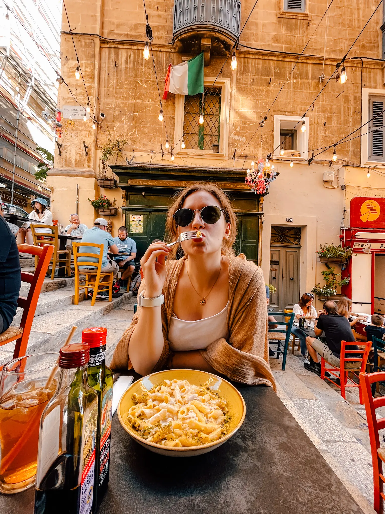 A girl eats pasta with a full plate in front of her, sitting in a colourful street with hanging lights in Valletta Malta.