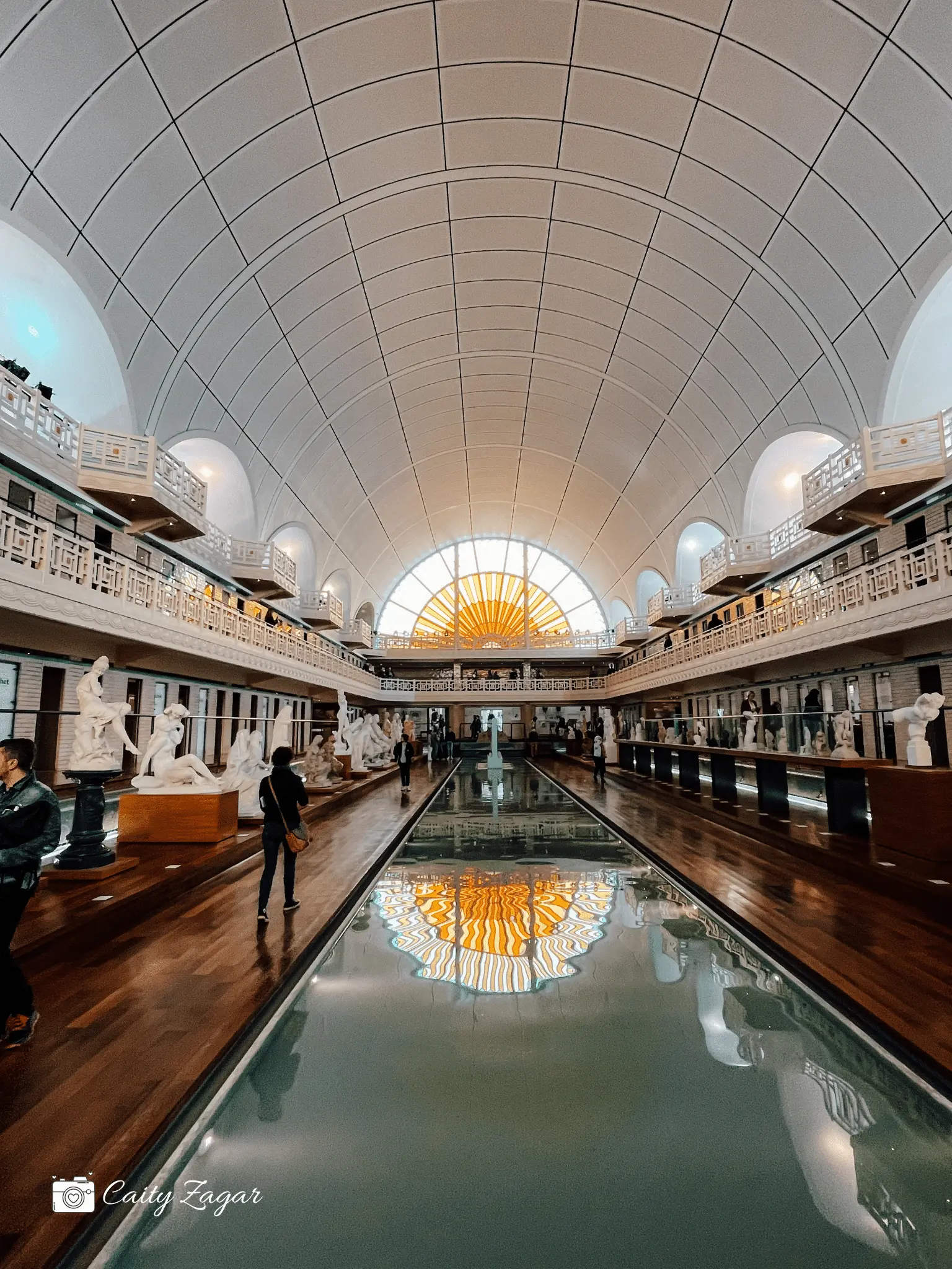 A semi-circle stained glass window sits at the end of a long swimming pool in La Piscine Musem in Roubaix. On either side of the pool are stages with sculptures and art deco balconies. 