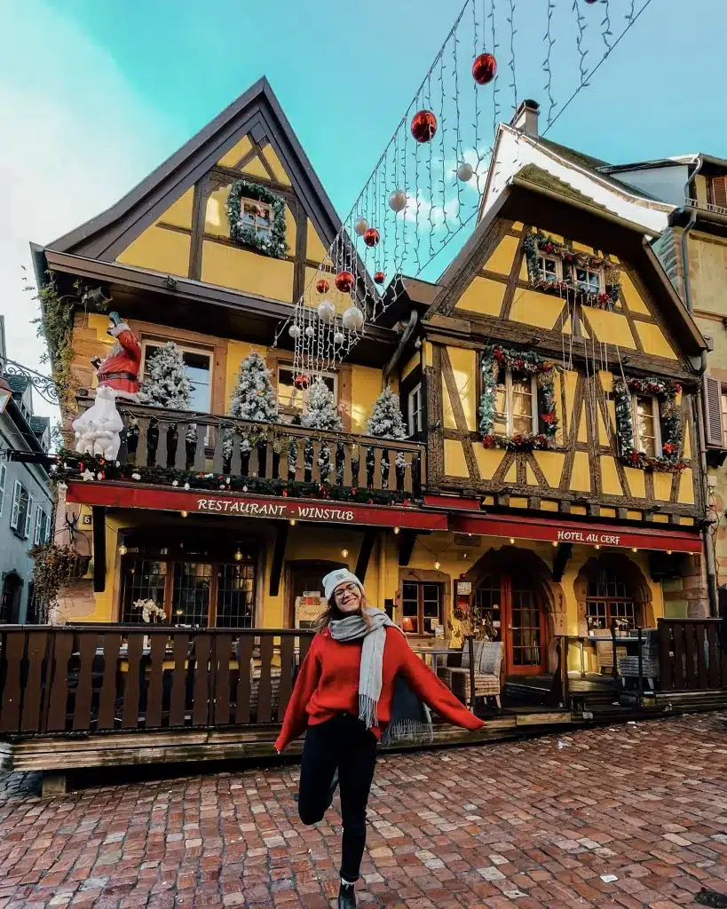 a girl smiling in front of a beautiful half-timbered yellow house covered in Christmas decorations in Alsace