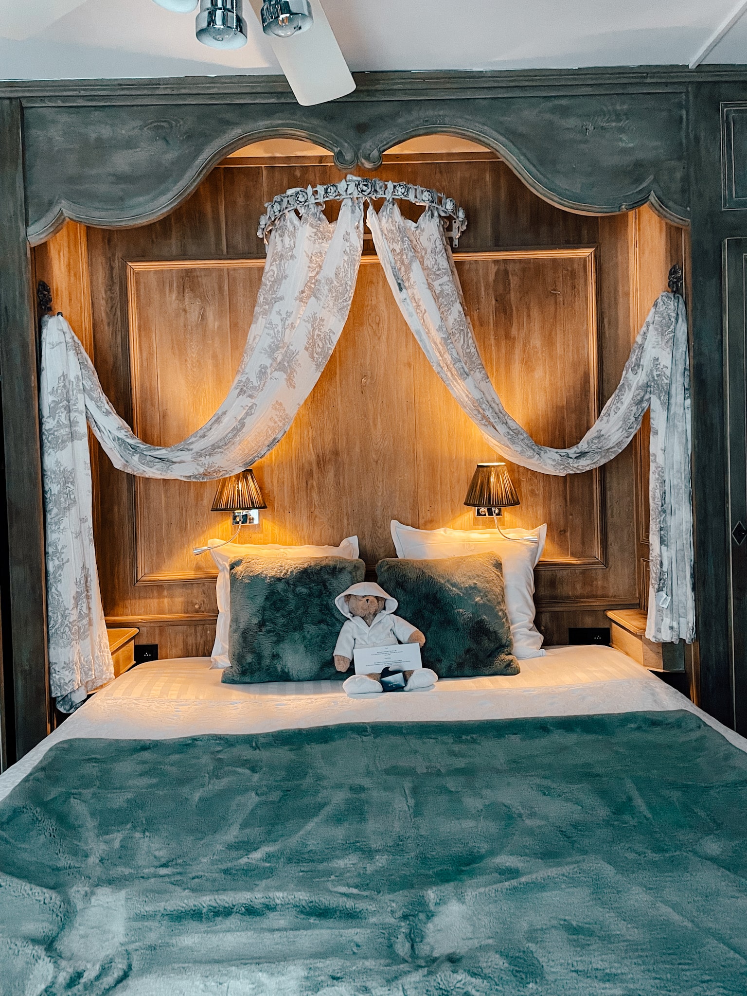 A perfectly set bed with a teddy bear sitting in front of the pillows. Above the bed hands 2 drapes and there are lights and other decorative features above the bed.